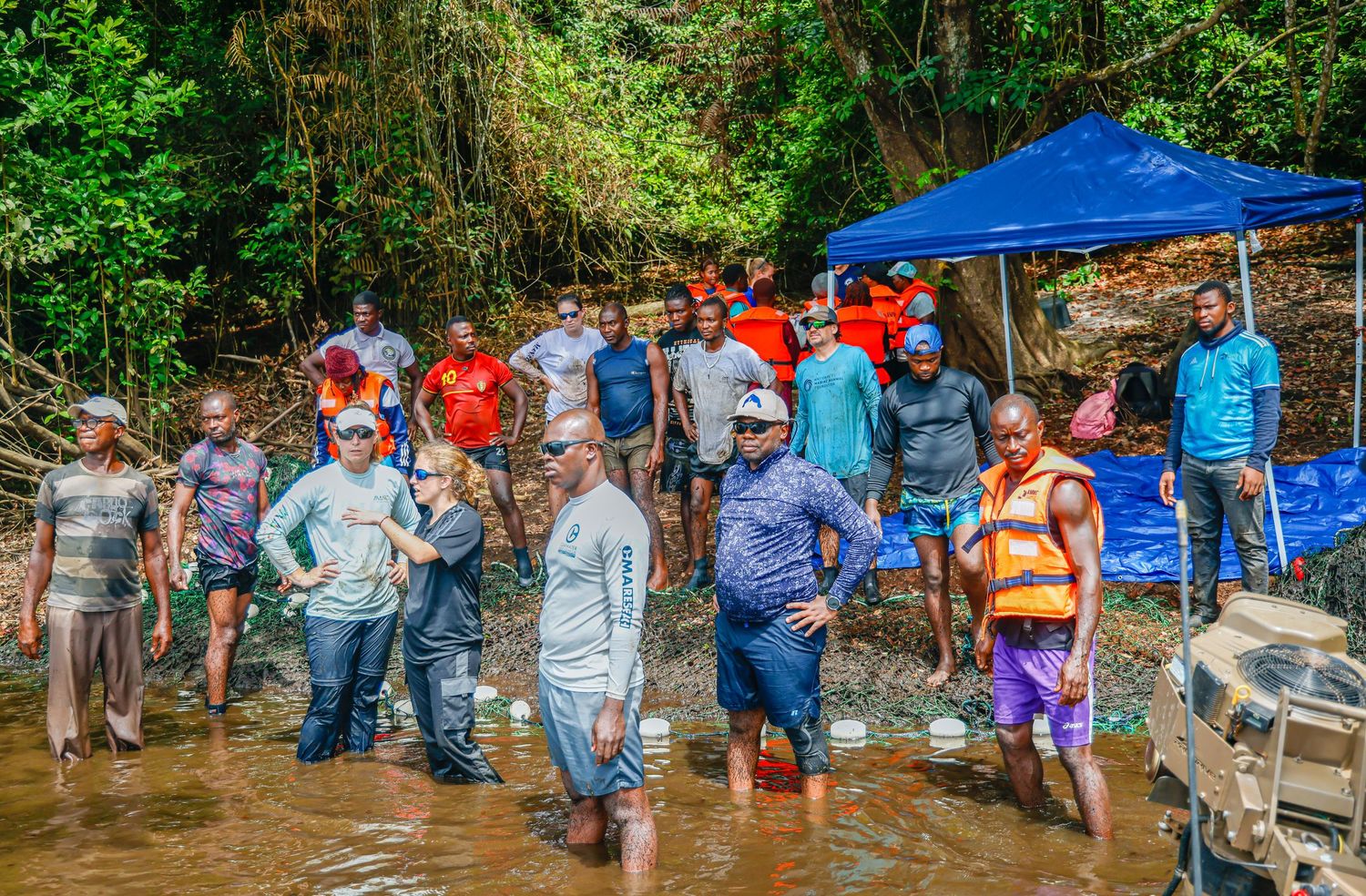 Group of people standing in a shallow river near a blue tarp canopy in a tropical forest setting.