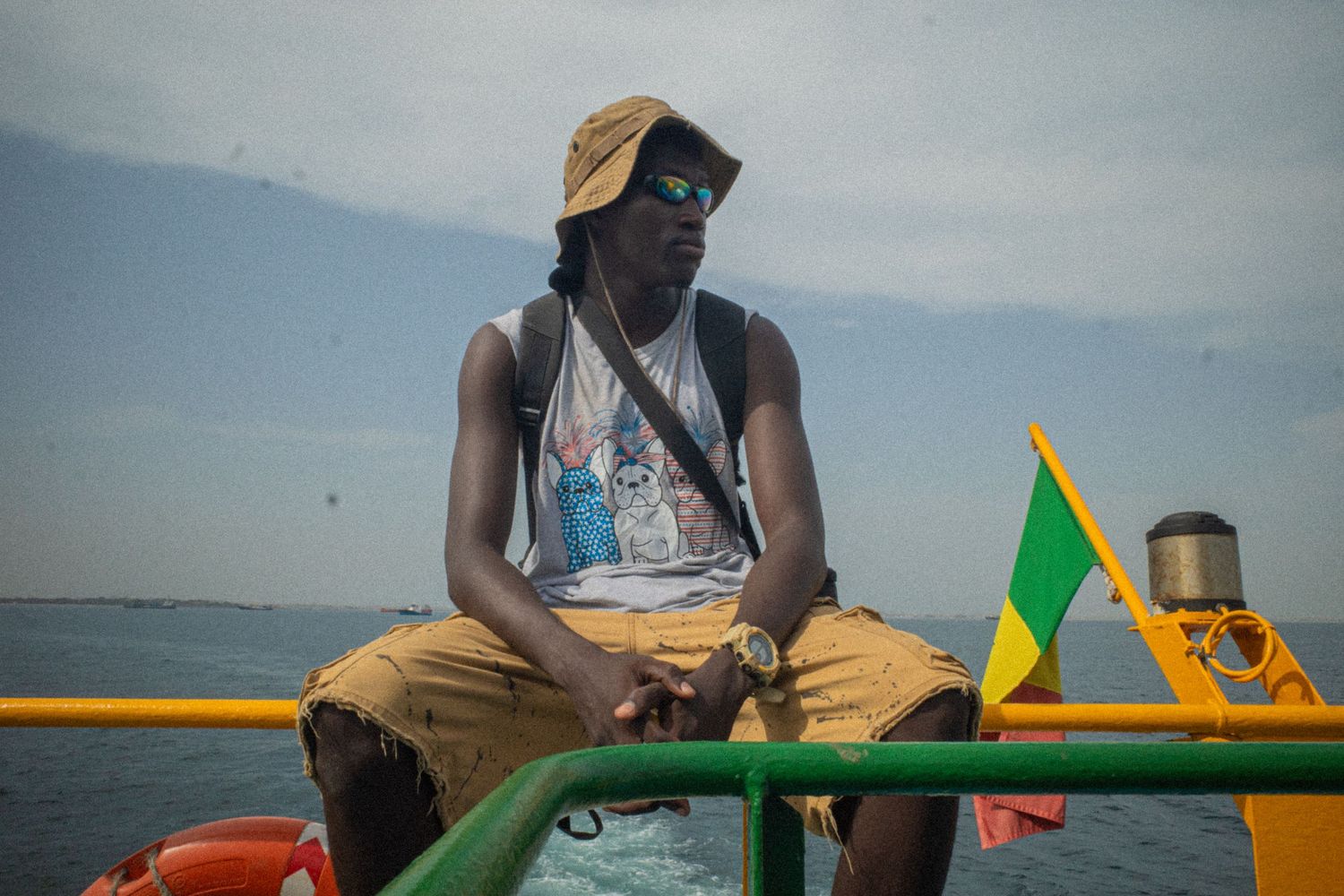 Young African man in sunglasses sitting on colorful ferry boat with Senegal flag visible.