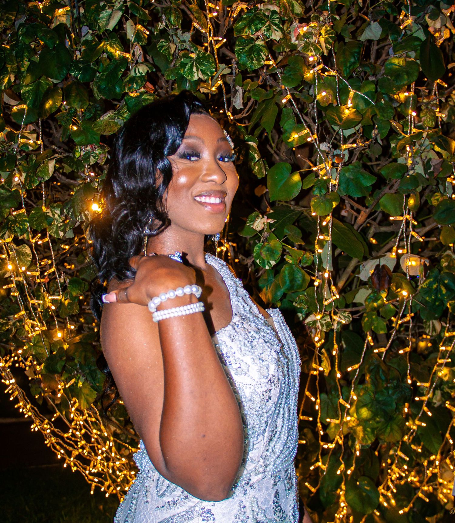 Woman in white lace dress smiling beside ivy wall adorned with fairy lights at night.