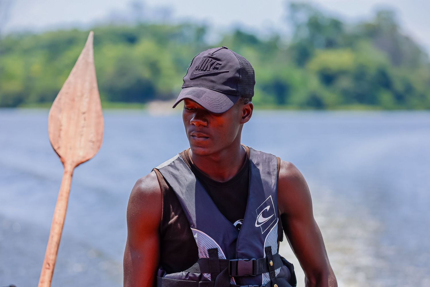A young man holding a wooden paddle stands near a river wearing a Nike cap.