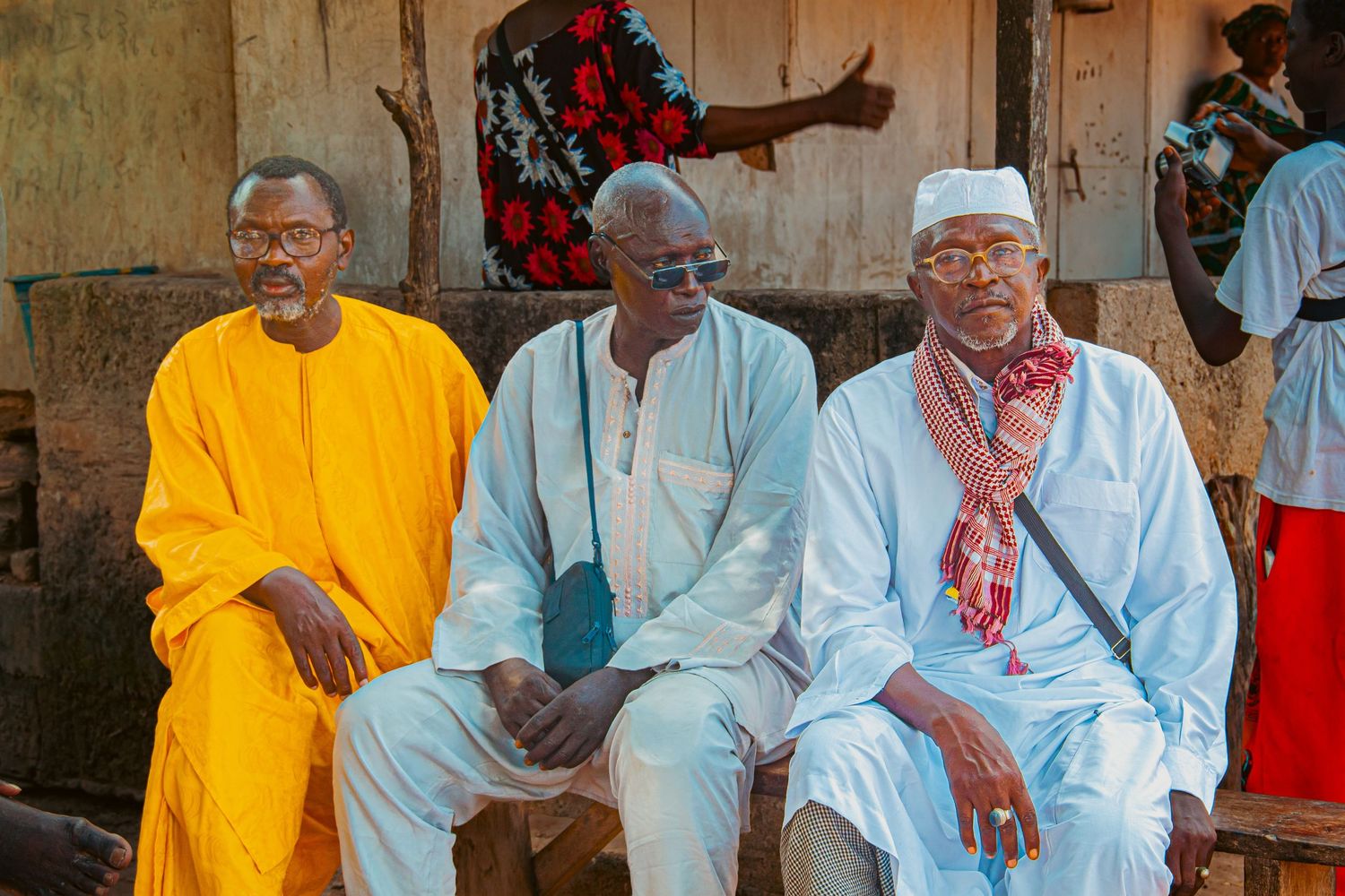 Three elderly African men in colorful traditional clothing sitting together outdoors.