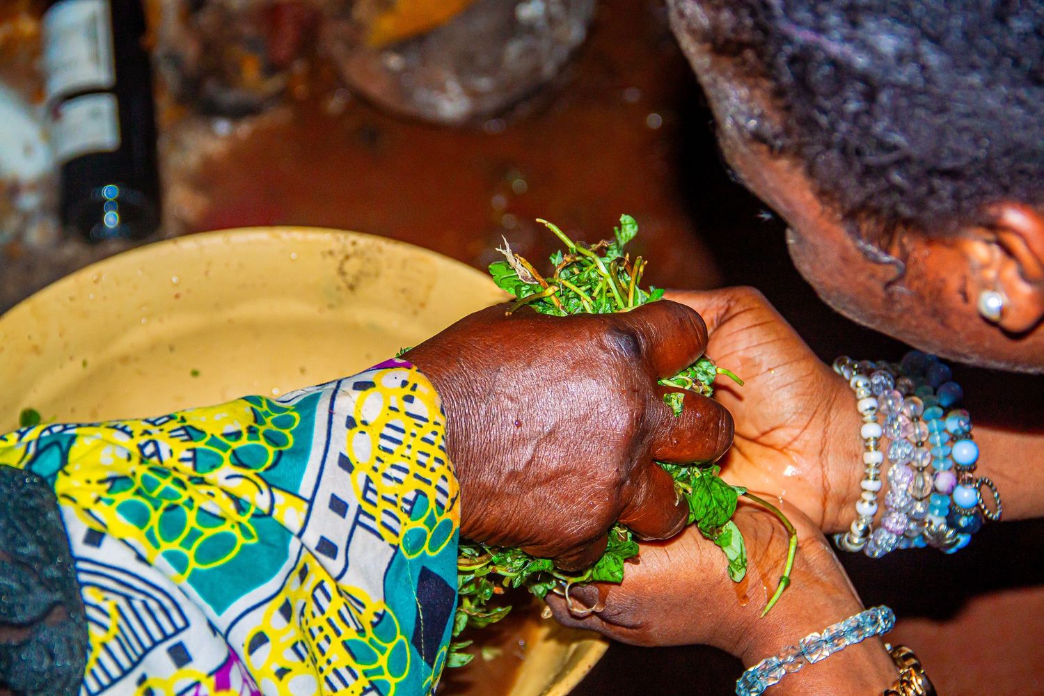 Hands adorned with bracelets holding green herbs over a bowl in African ritual ceremony.
