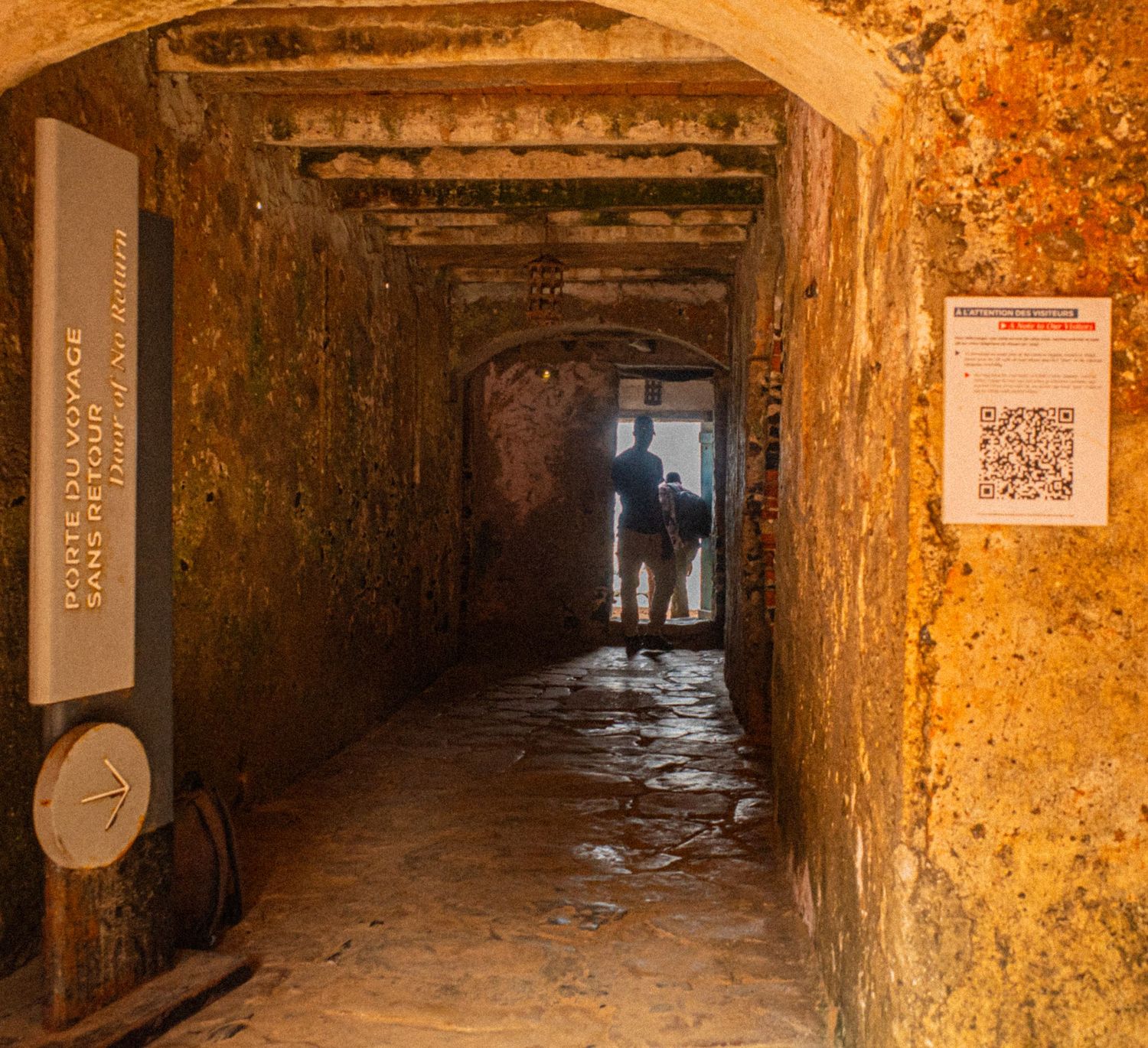 A dark narrow stone corridor leads to a bright doorway with two silhouetted figures inside.