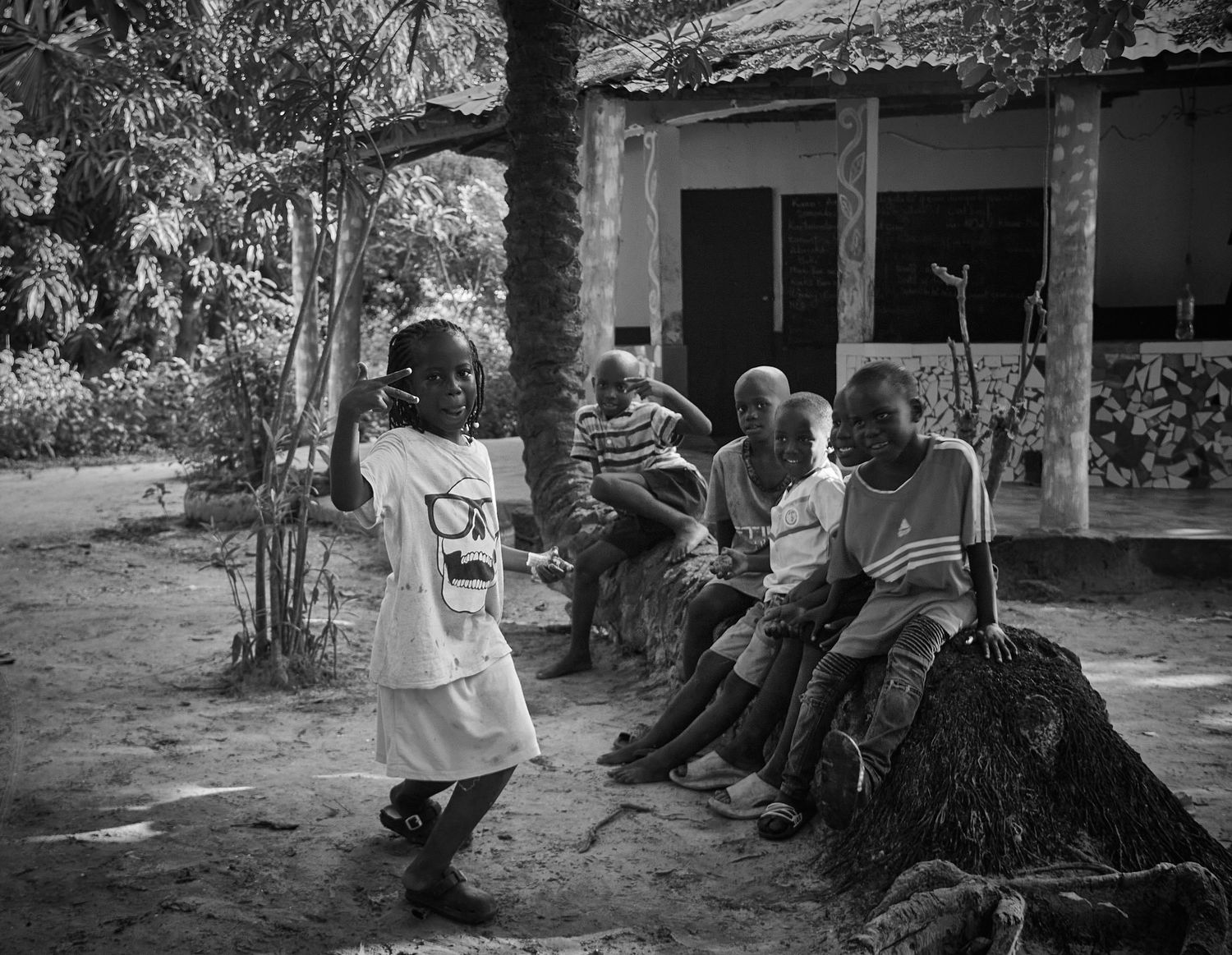 A young African girl in white dress dances before a group of seated elders in a black and white photo.