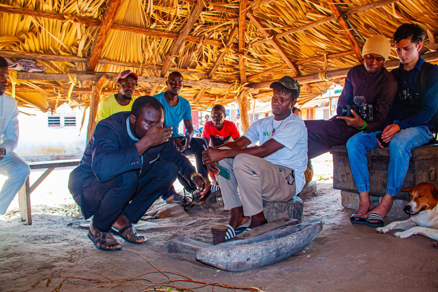 People gathered under thatched shelter in African village watching a traditional craft demonstration.