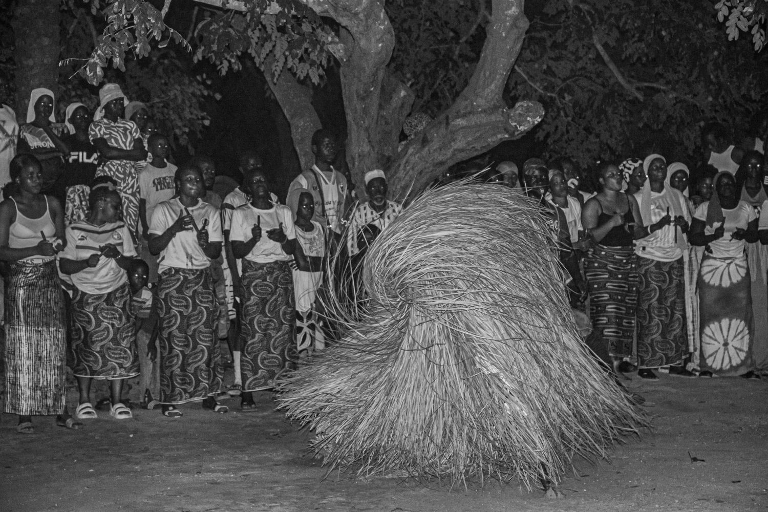 Black and white photo of a grass-costumed dancer performing at a traditional African nighttime ceremony.