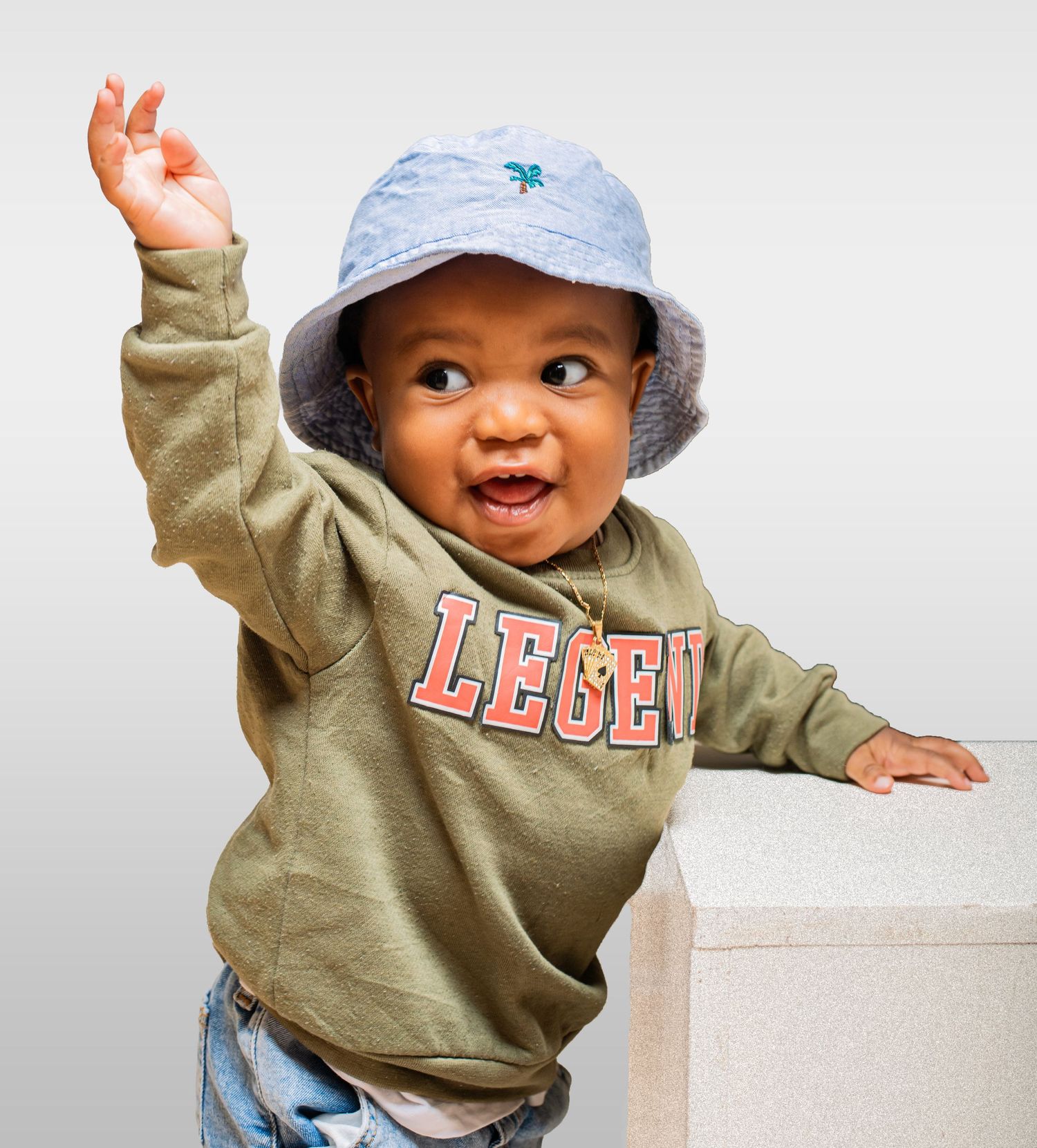 Happy baby in a Legend sweatshirt and bucket hat raising one hand playfully.