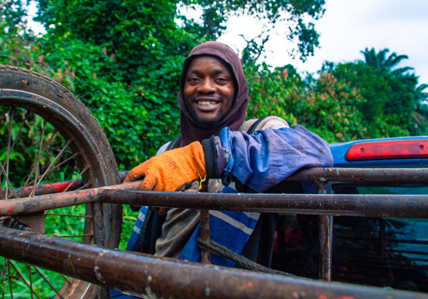 Smiling African man in a blue jacket and orange gloves leaning on a rusty farm vehicle outdoors.