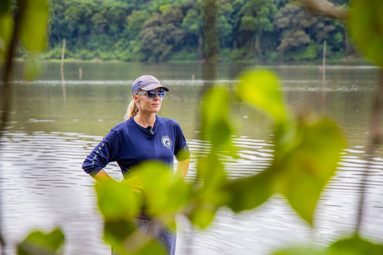 Woman in navy blue long-sleeve shirt standing near a river surrounded by green foliage.