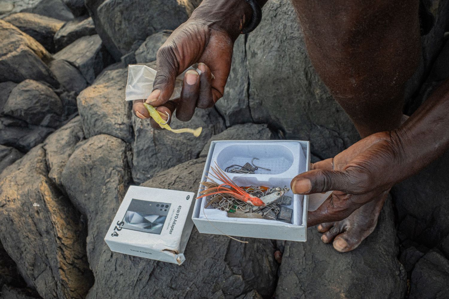 Hands of an African fisherman holding fishing lures and tackle box on rocky shore.