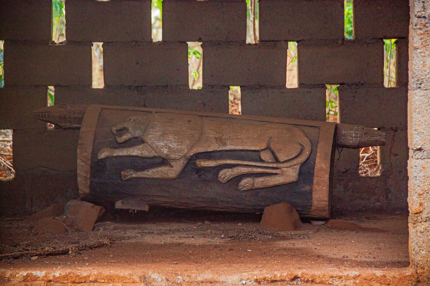 Ancient carved wooden drum with animal relief sculpture displayed inside traditional African mud brick structure.