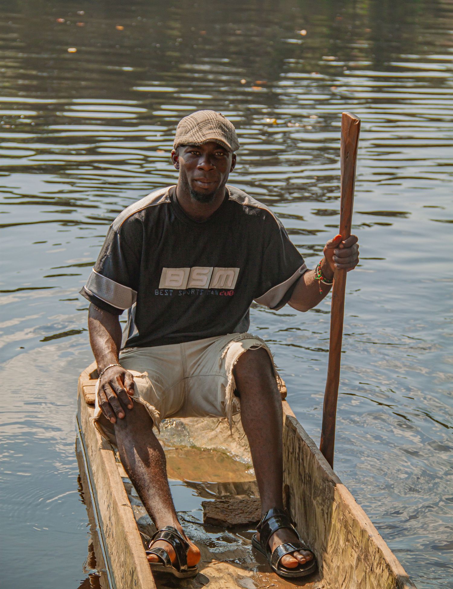 A man sits in a wooden canoe on a calm river, holding a paddle, wearing a cap and casual clothes.