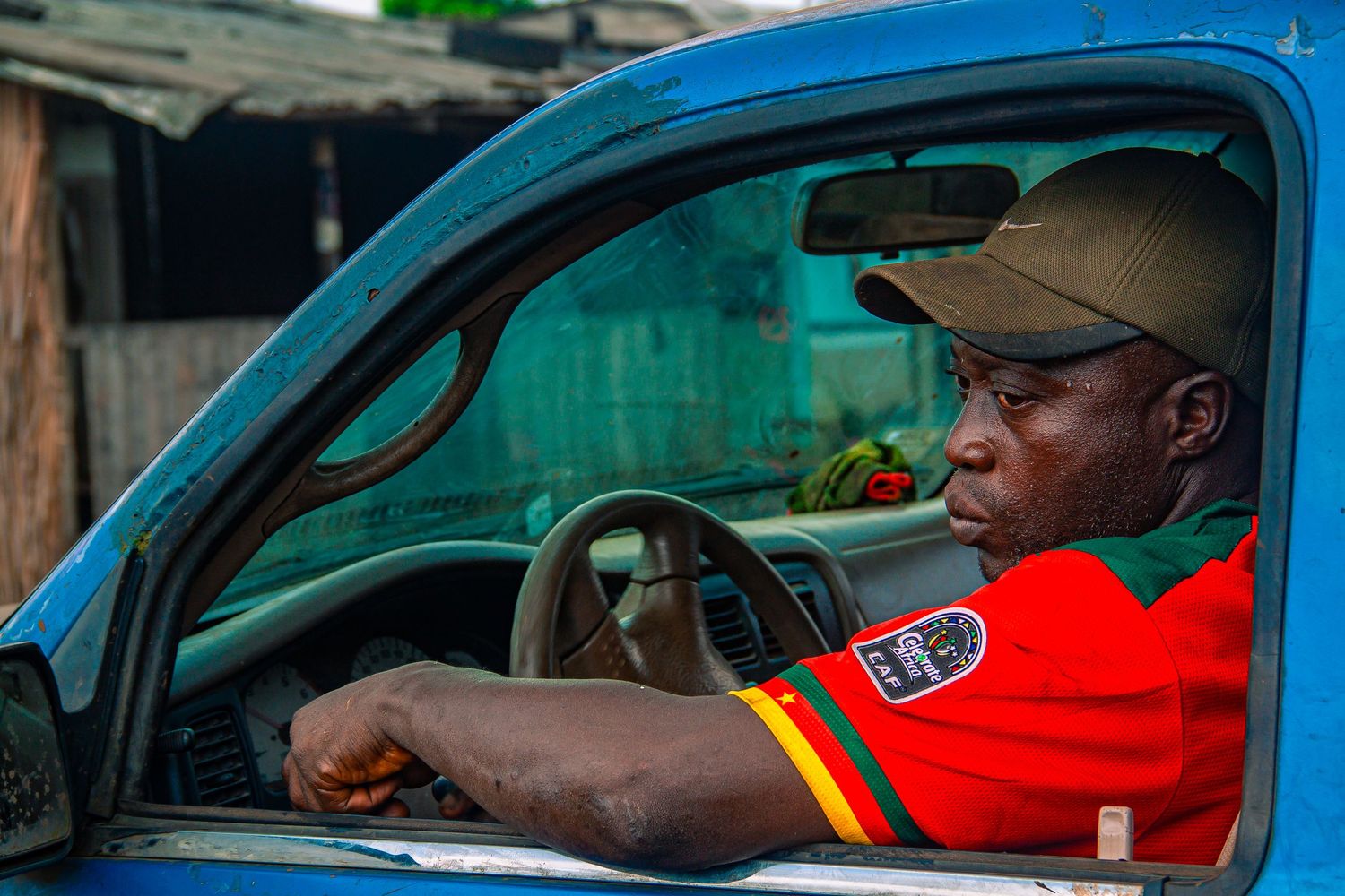 An African man in a red shirt and cap sits behind the wheel of a blue vehicle.