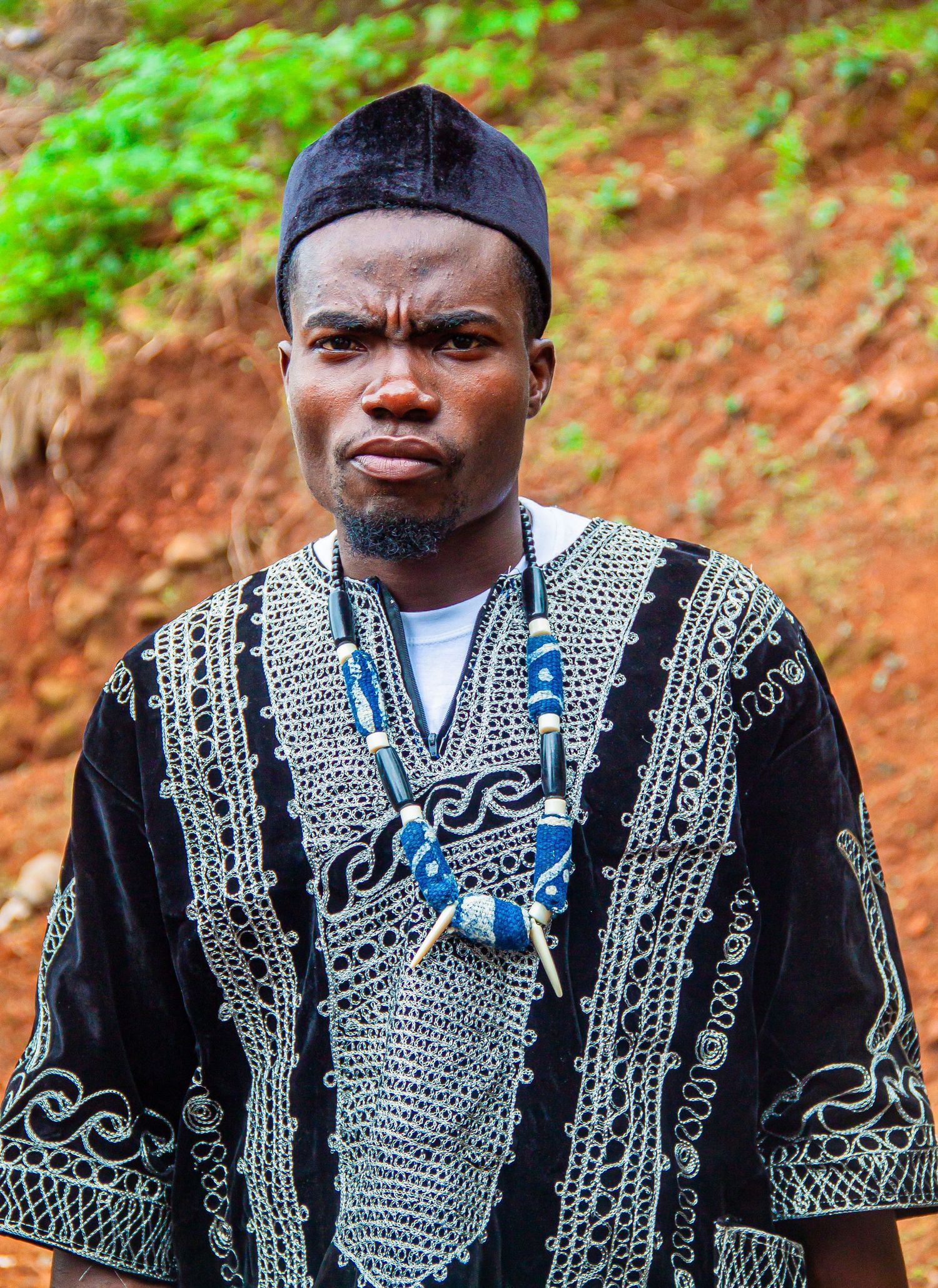An African man in traditional black embroidered dashiki and blue beaded necklace stands outdoors.