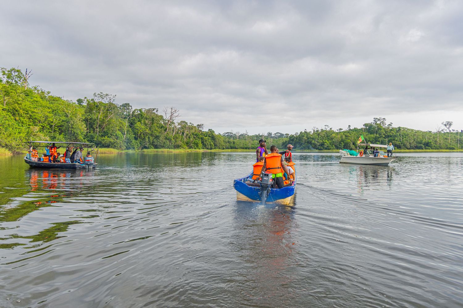 Small boats carrying passengers navigate a calm river surrounded by lush green tropical vegetation.
