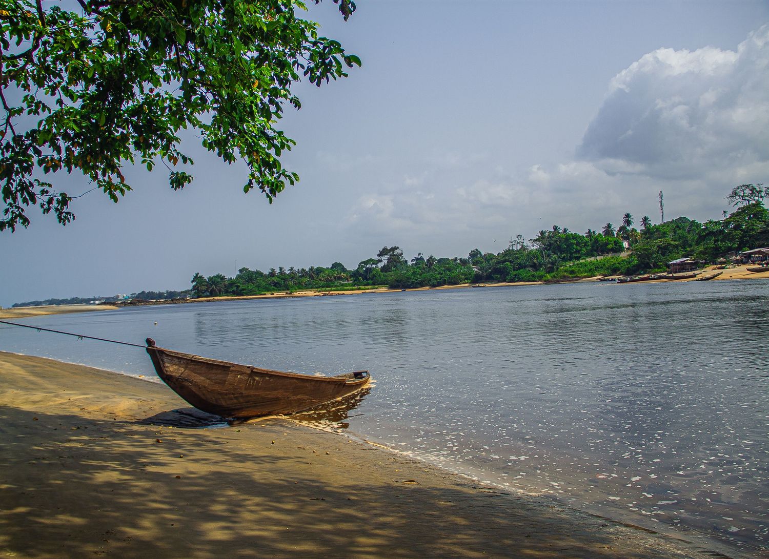 A wooden boat moored on a sandy riverbank with lush green trees and calm waters in the background.