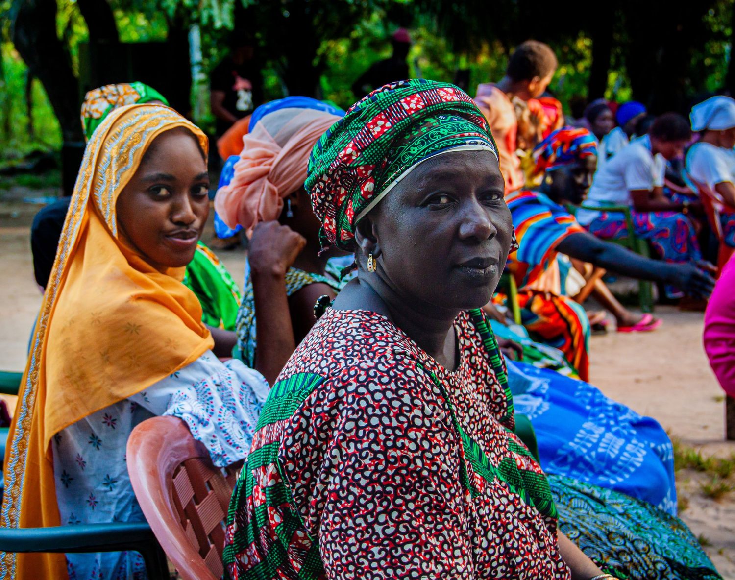 African women in colorful traditional headscarves and dresses seated outdoors at a gathering.