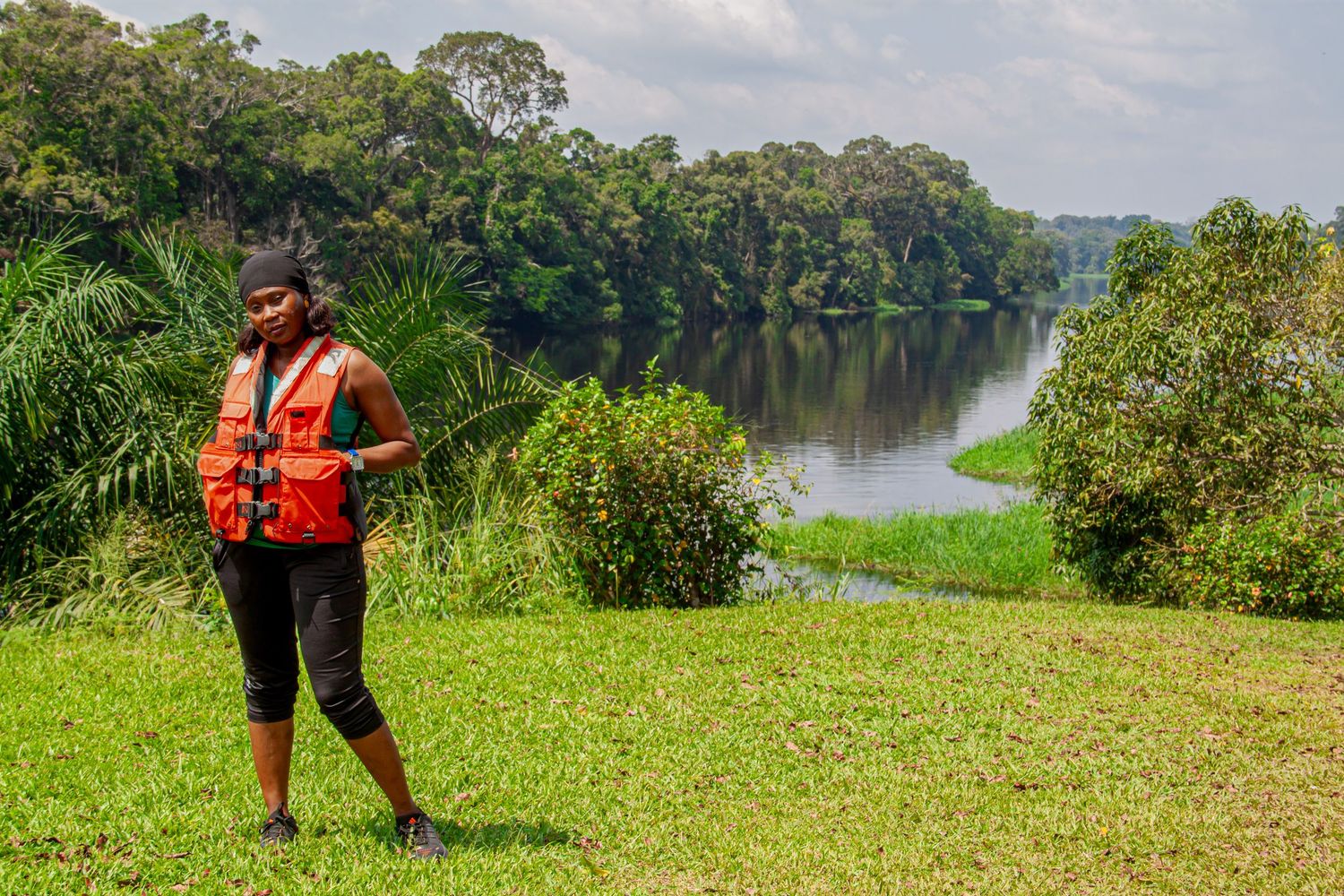 Woman in orange safety vest standing by a calm river in lush forest.