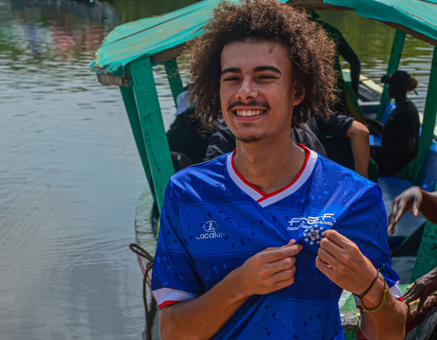 Smiling young man with curly hair in blue soccer jersey holding item on a boat.