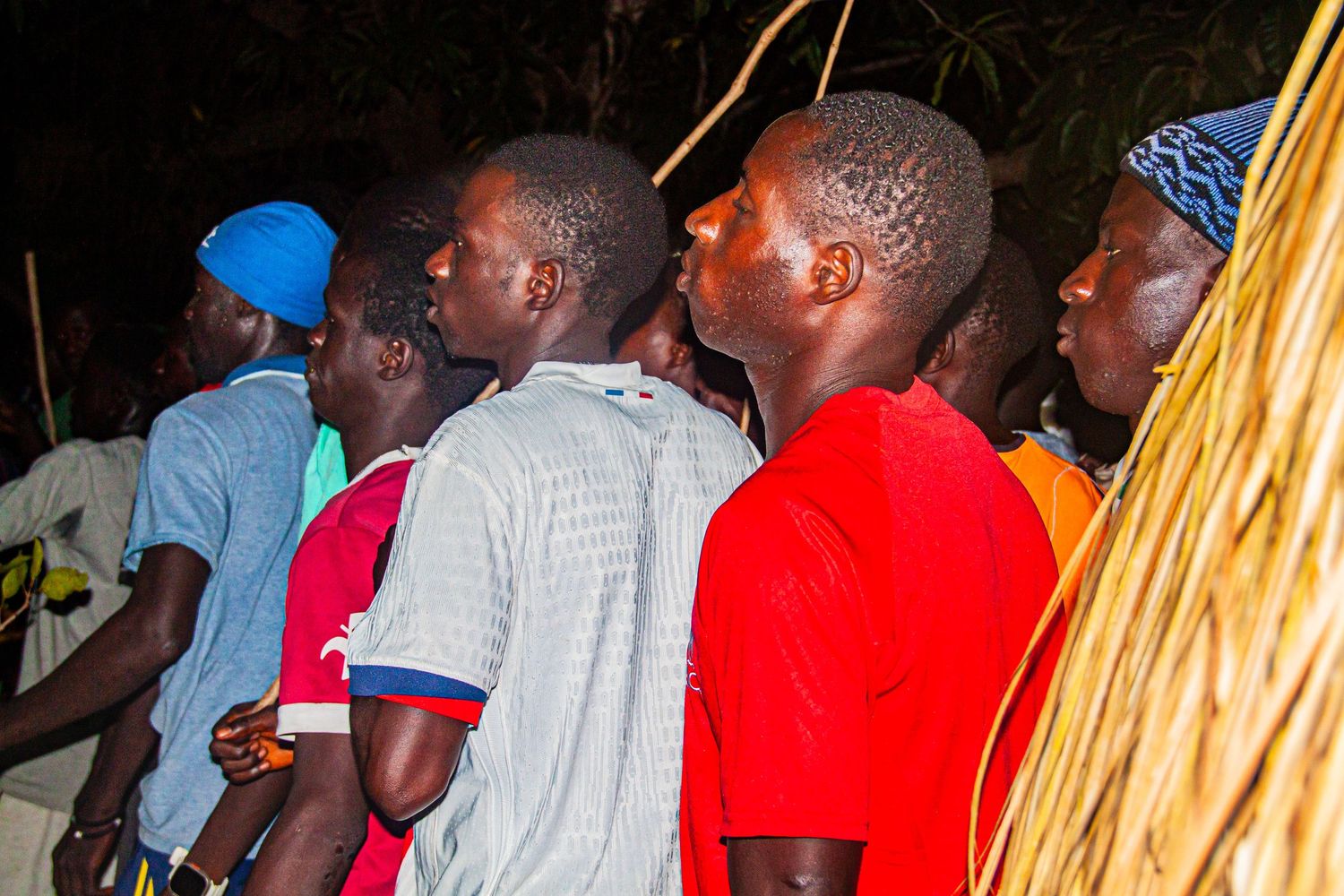 Group of young African men watching a traditional grass costume performance attentively at a night event.