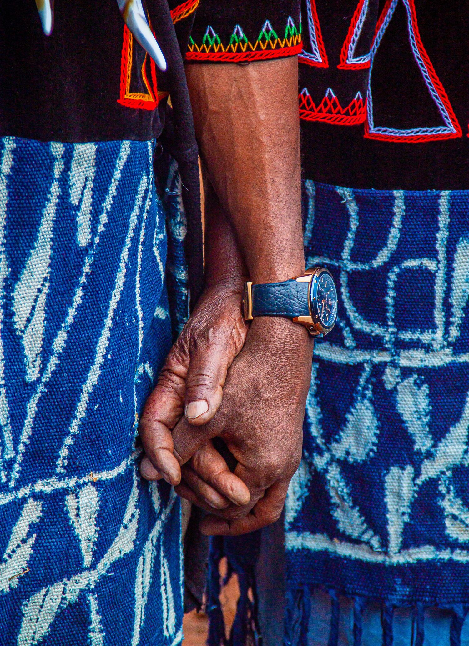 Close-up of hands wearing a watch and traditional blue Ndop patterned fabric at a cultural event.