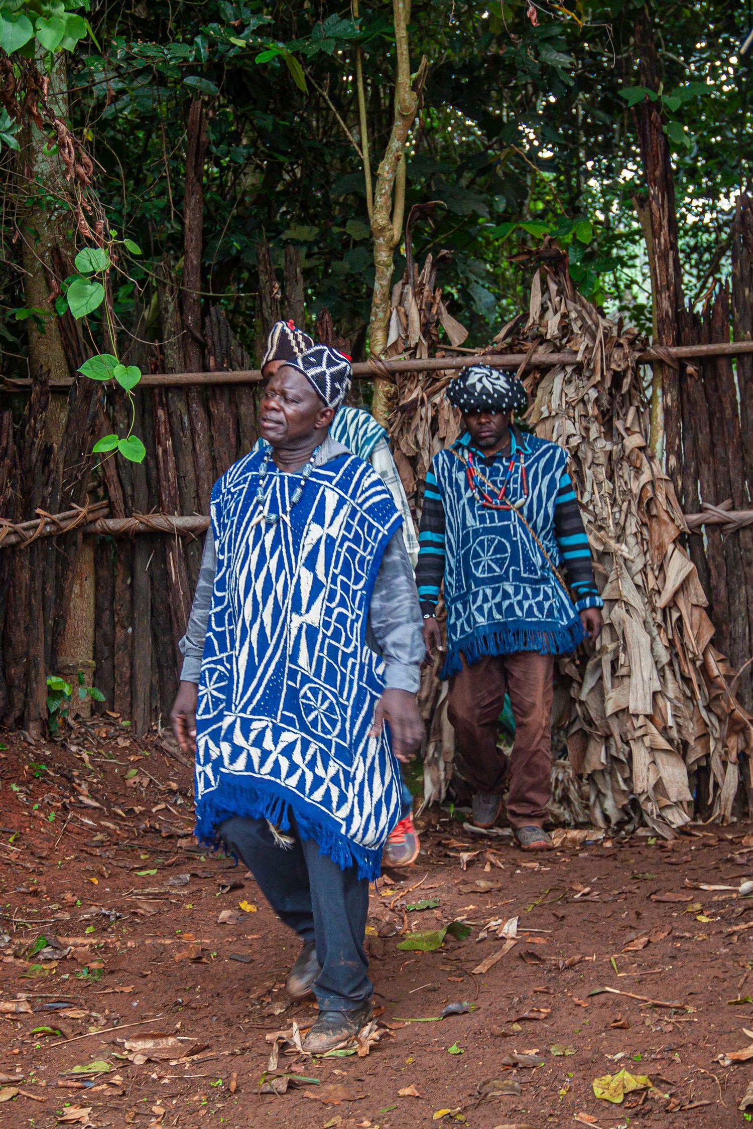 Two African men in traditional blue and white patterned Cameroonian attire near a bamboo fence.