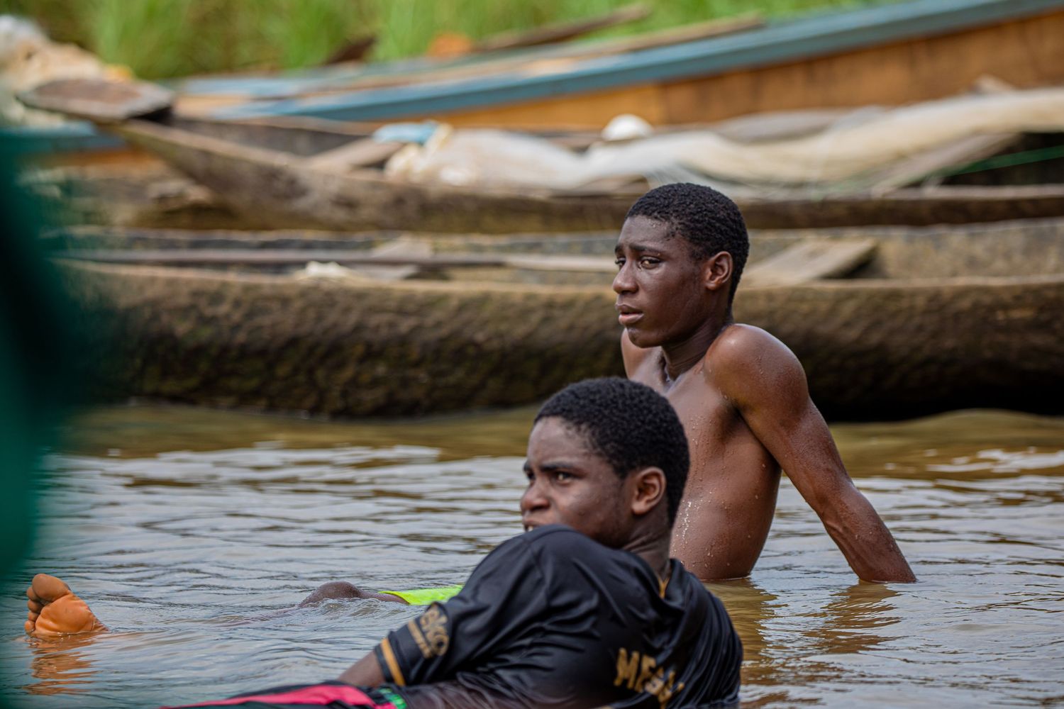 Two young African boys wading in a river near wooden boats in a tropical waterway setting.