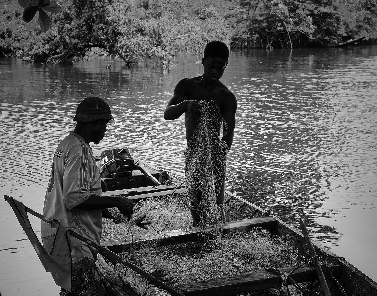 Two fishermen working with nets on a wooden boat in a calm river, black and white photo.