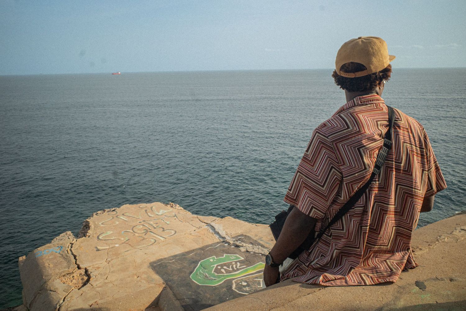 Man in traditional African attire sitting on a sea wall gazing at the ocean.