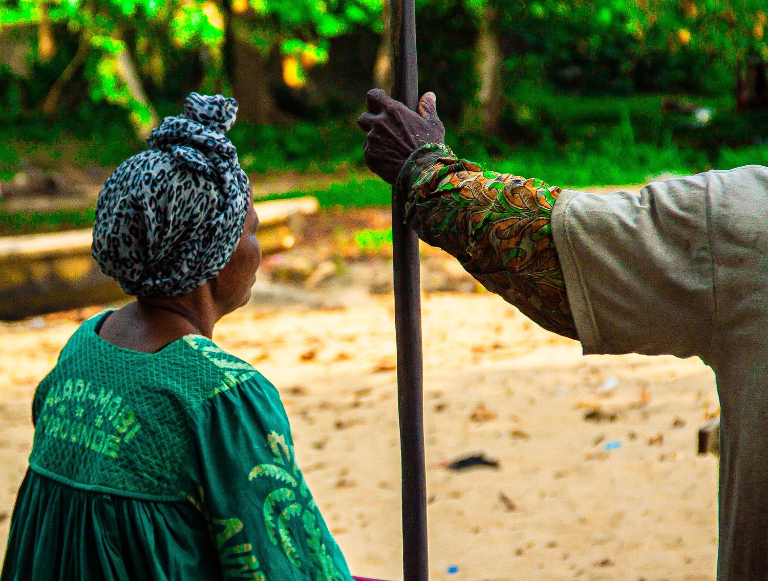 Young child in green outfit with head wrap looks up at an adult holding a wooden staff outdoors.