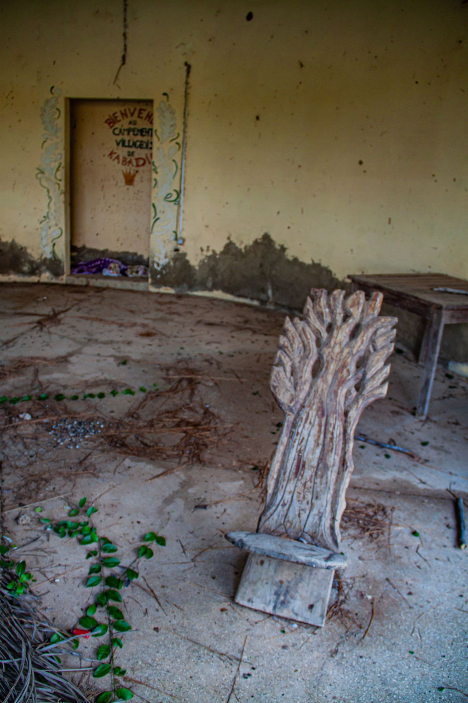 Abandoned room with rustic wooden carved chair on dirt floor and faded painted door in background.
