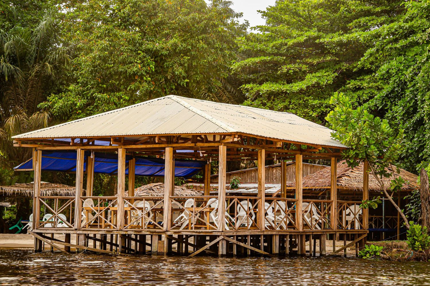 A wooden stilt pavilion restaurant over calm water surrounded by lush tropical rainforest trees.