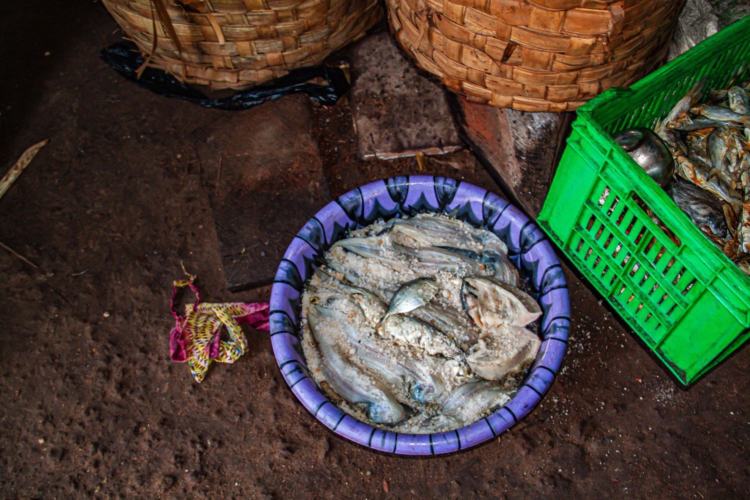 Fresh fish displayed in a purple bowl on a wet market floor beside wicker baskets and a green crate.