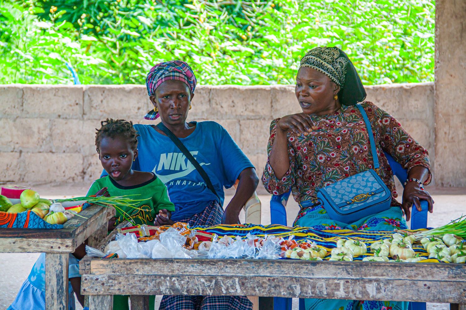 Three African women selling fresh vegetables and produce at an outdoor market stall.