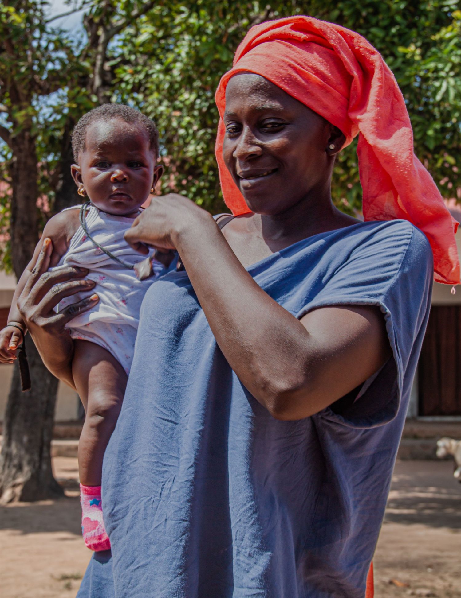 Smiling African mother in blue dress and red headscarf holding her baby outdoors.