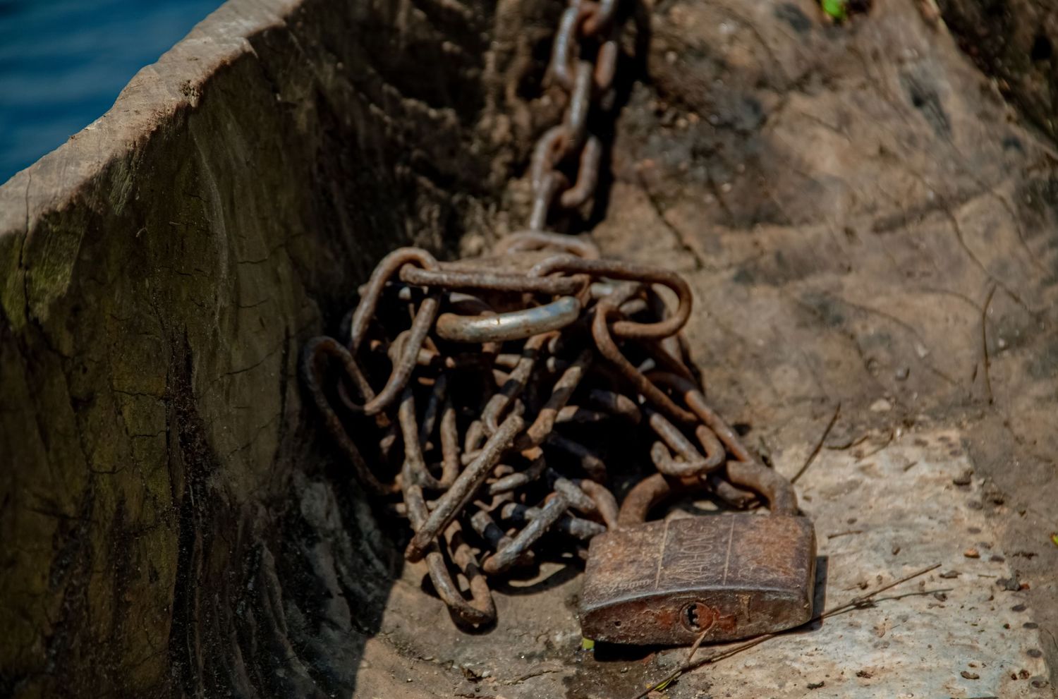 Rusty iron chains and padlock resting on weathered stone surface near water in sunlight.