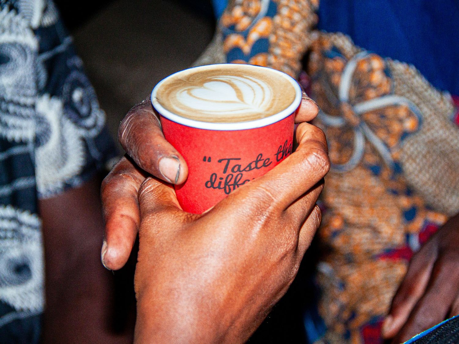 Hands holding a red cup of latte art coffee with colorful African fabric in background.