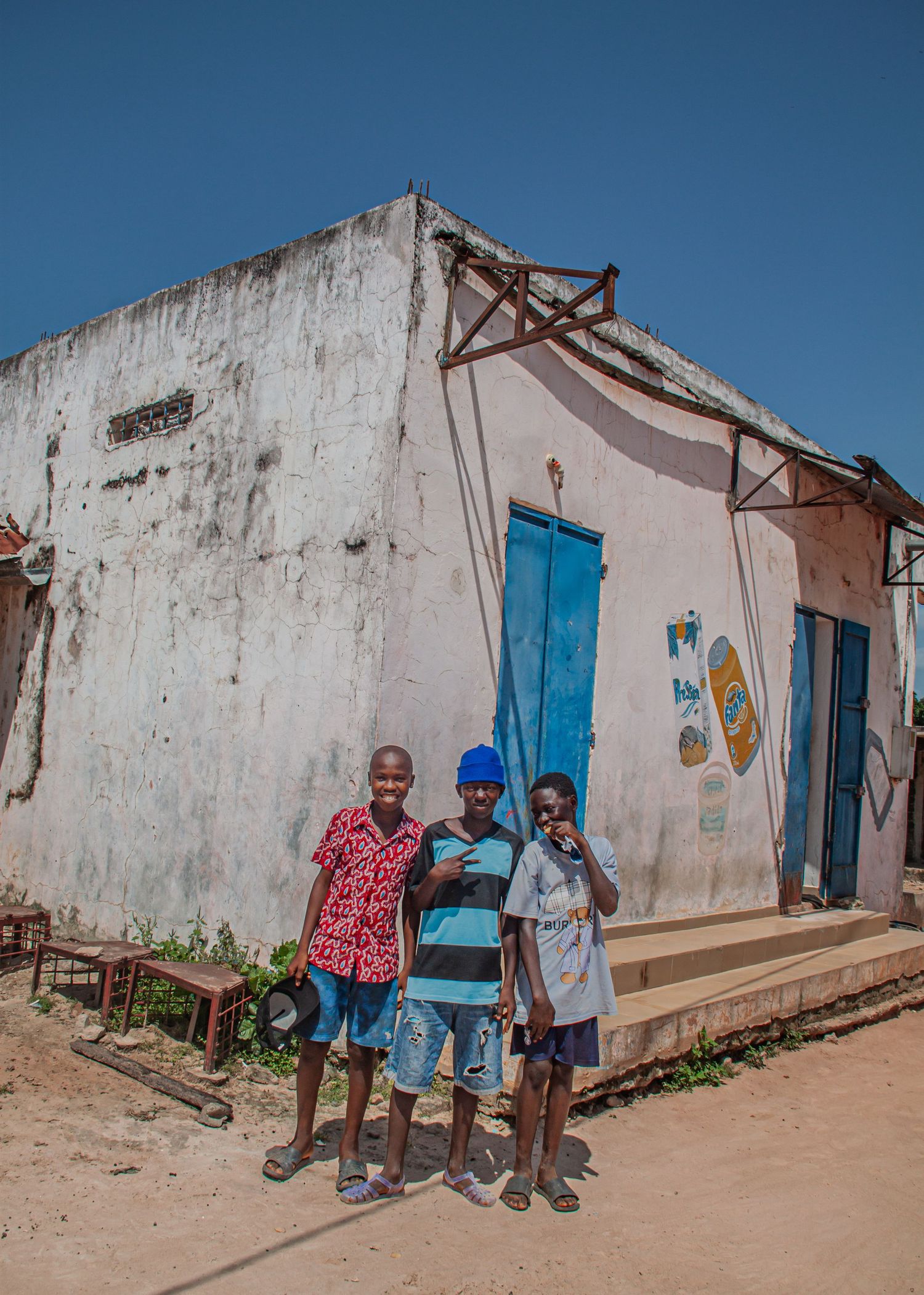 Three young African boys pose smiling in front of a weathered white building with a blue door.