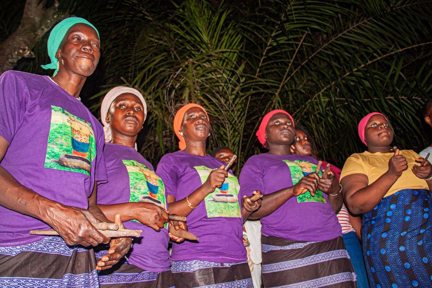 Group of African women in matching purple t-shirts performing a traditional dance outdoors at night.
