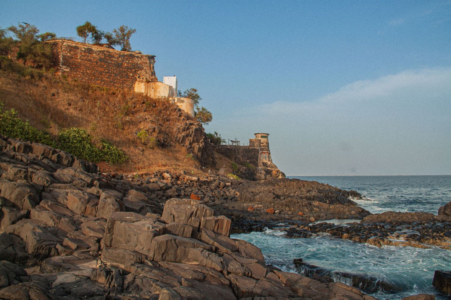 Ancient stone fortress ruins on rugged coastal cliffs overlooking a blue ocean with crashing waves under clear sky.