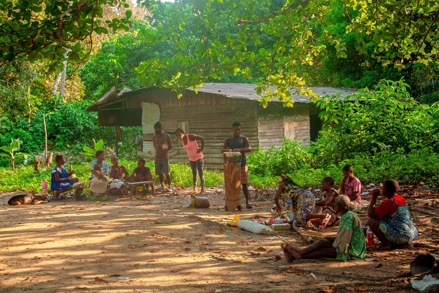 African villagers gathered in a sunny clearing with a rustic wooden building and green trees.