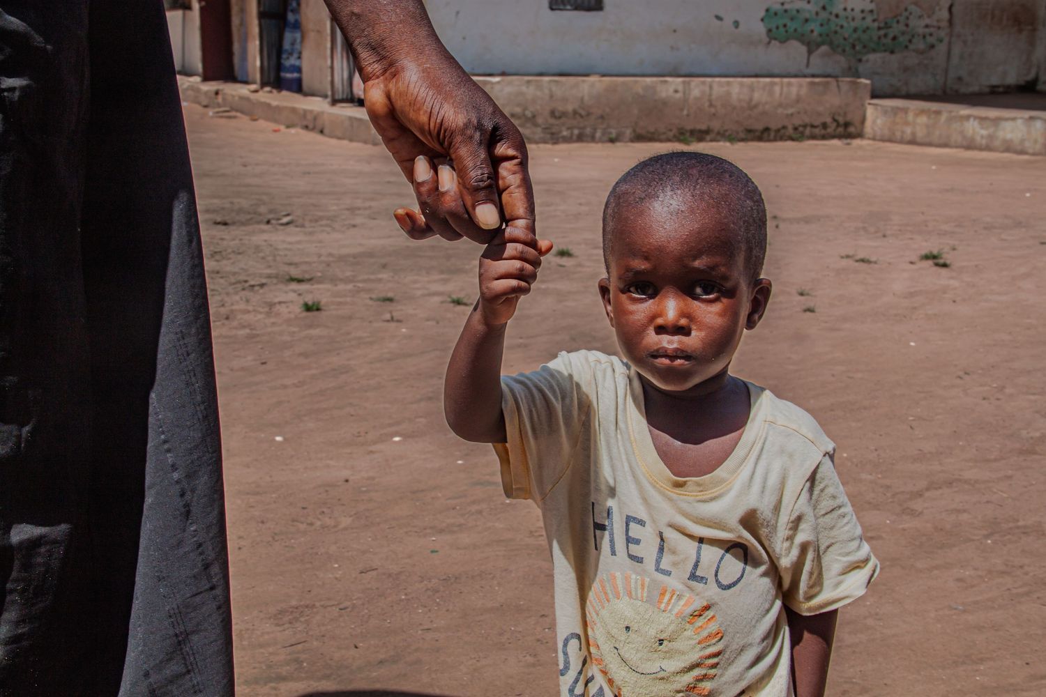 A young African child in a Hello Kitty shirt holds an adult's hand on a dusty village street.