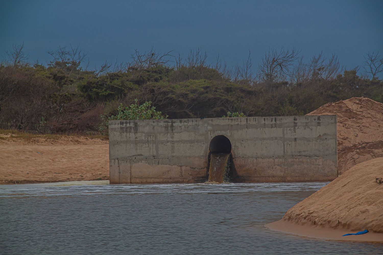 Concrete drainage tunnel outlet releasing water into a sandy riverbank landscape under a stormy blue sky.