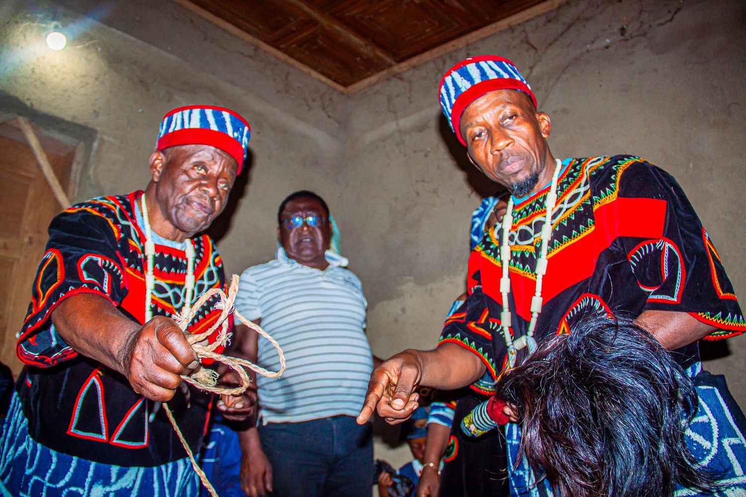 Two men in traditional African attire with colorful beaded hats perform a cultural ceremony indoors.