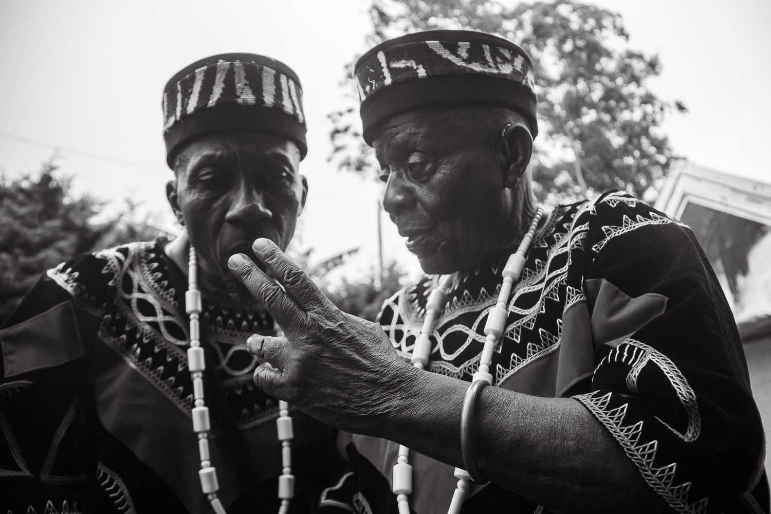 Two elderly African men in traditional embroidered attire share a conversation in black and white photography.