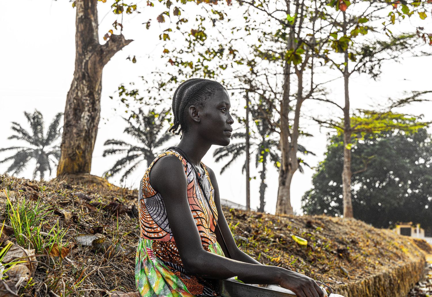 A young woman in a colorful dress sits pensively on a mound surrounded by trees in a rural landscape.