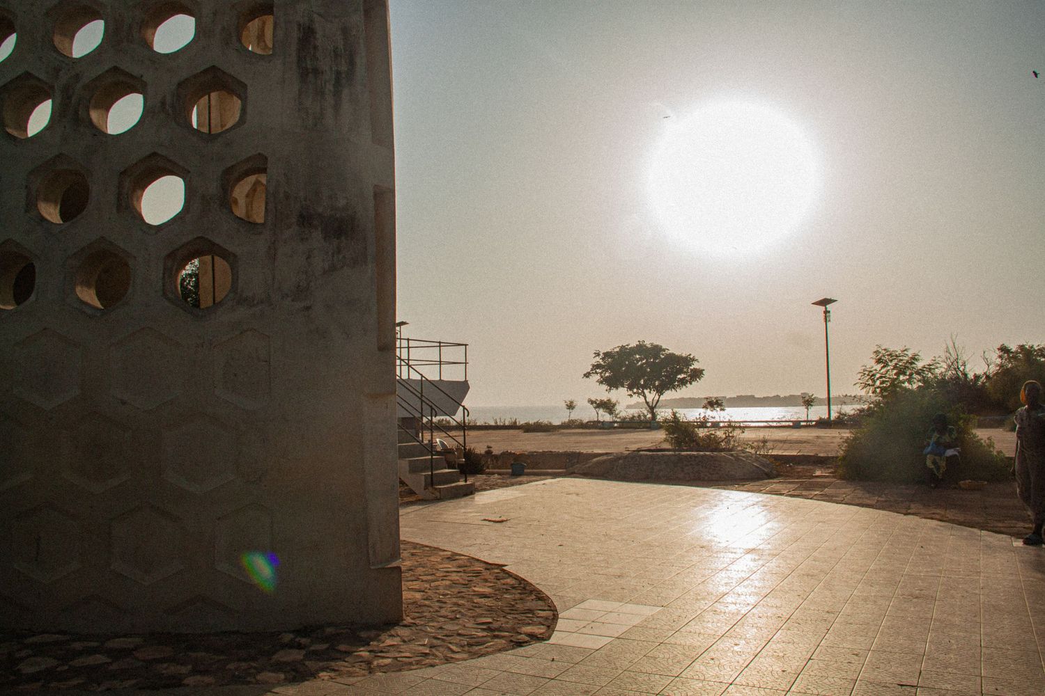 Bright sun flare over a sandy coastal plaza featuring a decorative geometric concrete wall and a lone tree at sunset.