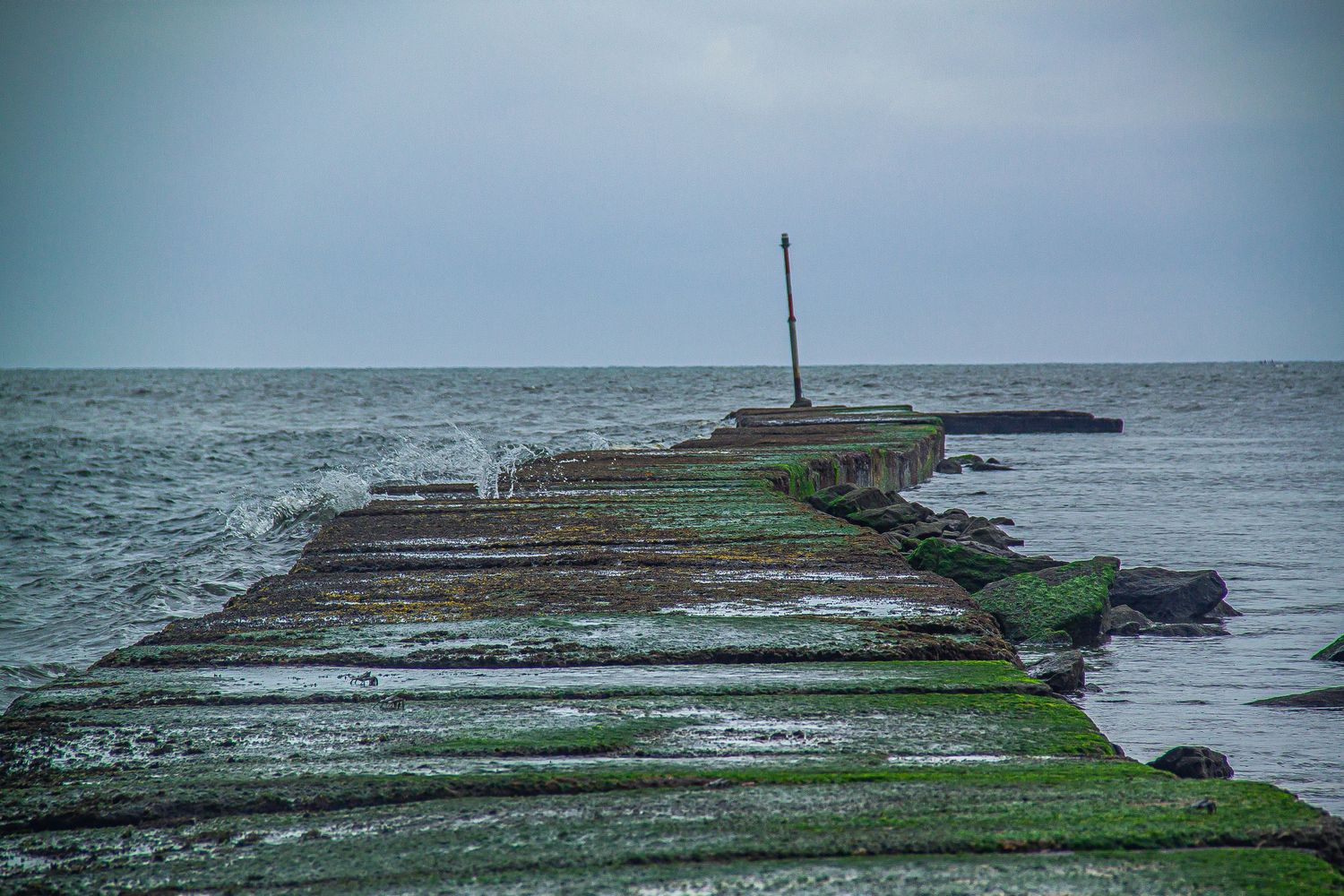 Algae-covered stone jetty stretching into a rough gray sea under cloudy skies.