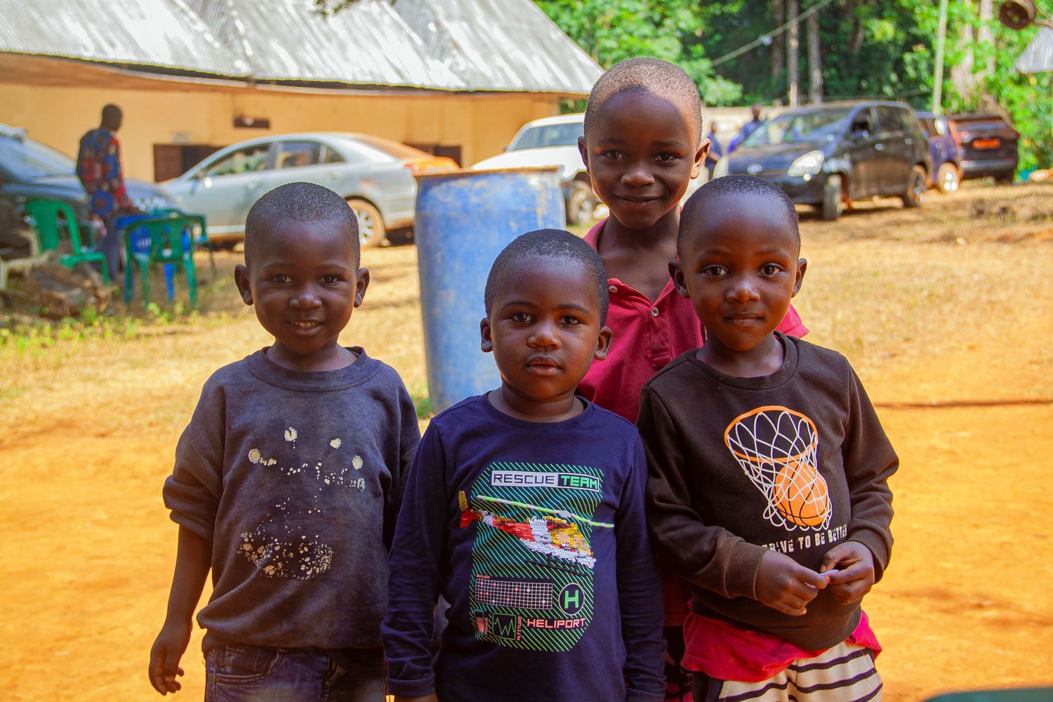 Four young African children smiling together on red dirt ground outside.