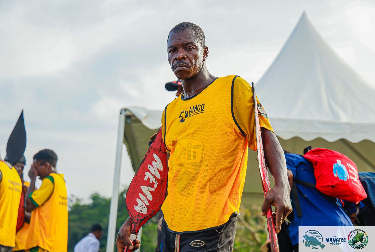 A man in a yellow AMCO jersey holds equipment at an outdoor sports event in Africa.